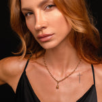 Woman with wavy, light brown hair in a black spaghetti strap top and gold necklace poses against a black background, her cross tattoo and Witch’s Bell Charm adding a touch of protective magick.