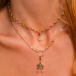 A close-up of a model's neck and collarbone wearing a layered gold necklace set. The set features a delicate chain choker with a small Witch's Knot charm and a longer chain with a gold ring and a small birdhouse-shaped pendant.