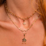 A close-up of a model's neck and collarbone wearing a layered gold necklace set. The set features a delicate chain choker with a small Witch's Knot charm and a longer chain with a gold ring and a small birdhouse-shaped pendant.