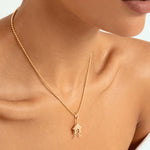 A close-up of a model wearing a delicate gold chain necklace with a small house-shaped pendant featuring a tiny figure inside, set against a clean white background.