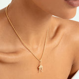 A close-up of a model wearing a delicate gold chain necklace with a small house-shaped pendant featuring a tiny figure inside, set against a clean white background.