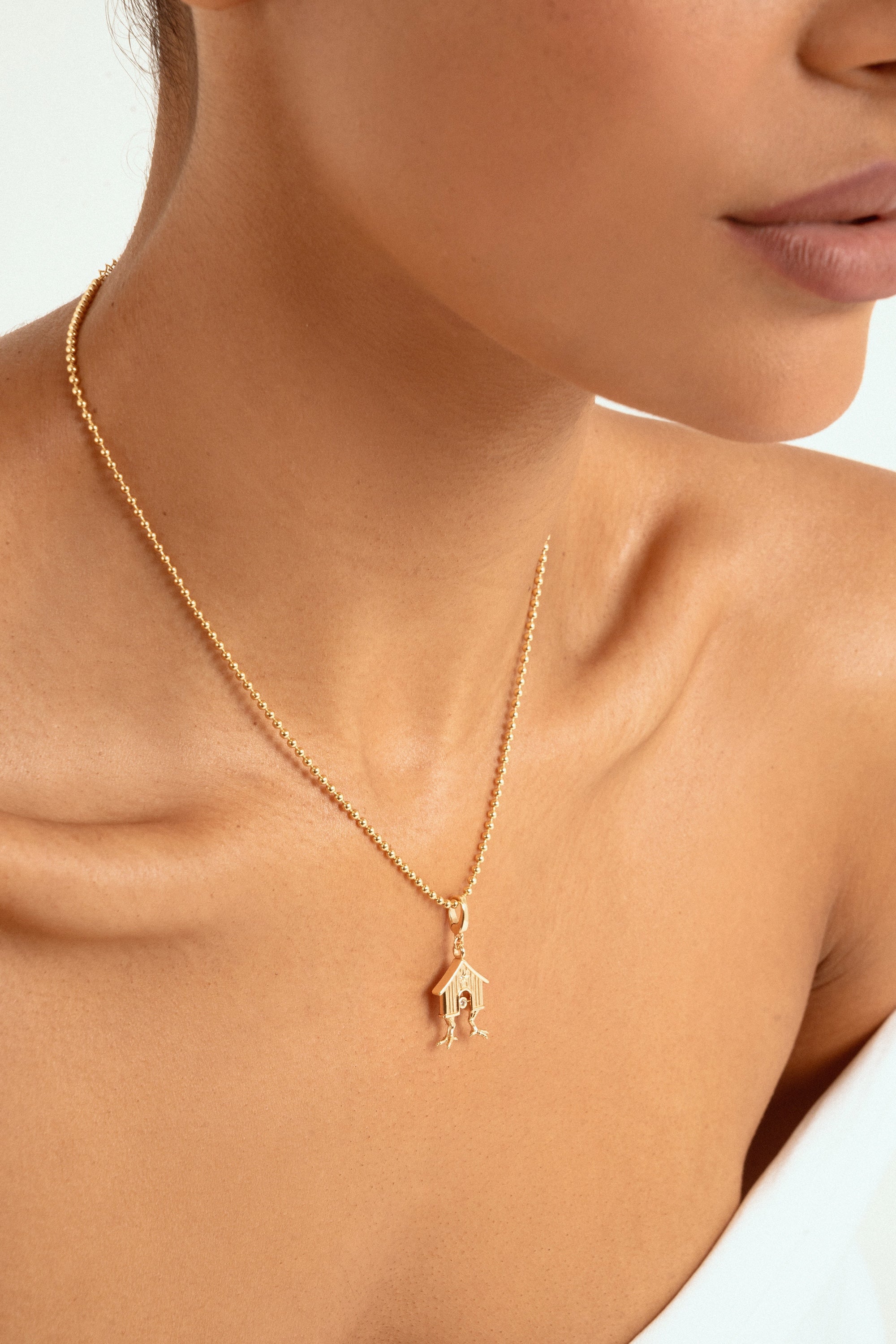 A close-up of a model wearing a delicate gold chain necklace with a small house-shaped pendant featuring a tiny figure inside, set against a clean white background.
