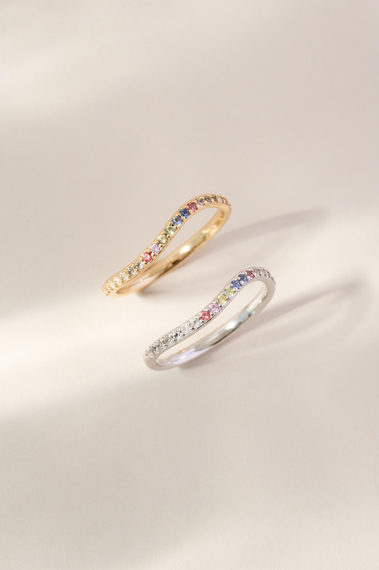 Two rings, one gold and one silver, with colorful gemstones on a light beige background.