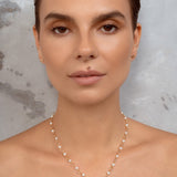 A close-up of a woman wearing a delicate gold necklace with small pearl-like beads and two pendants, a key and a caduceus symbol. against a textured gray background.