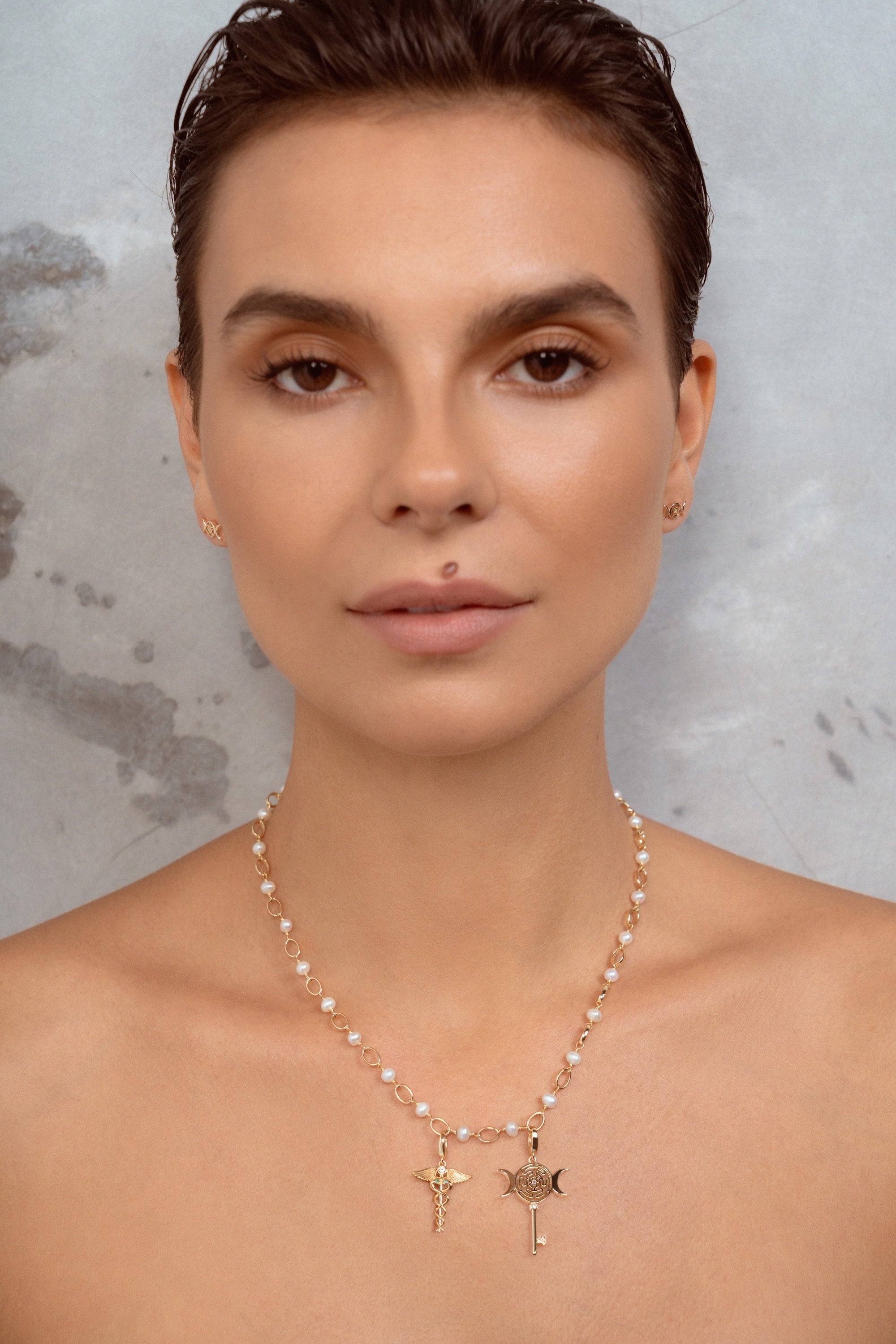A close-up of a woman wearing a delicate gold necklace with small pearl-like beads and two pendants, a key and a caduceus symbol. against a textured gray background.
