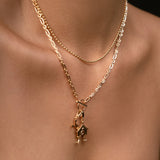 A close-up of a model's neck and chest wearing a layered gold necklace set. The top layer features a delicate gold beaded chain, while the bottom layer has a slightly thicker gold chain with a small pendant and a central house charm.
