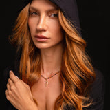 A woman with long, wavy red hair wears a black hooded top and the Preorder: Hades Charm (Ships end October 2026) against a black background, her expression neutral.