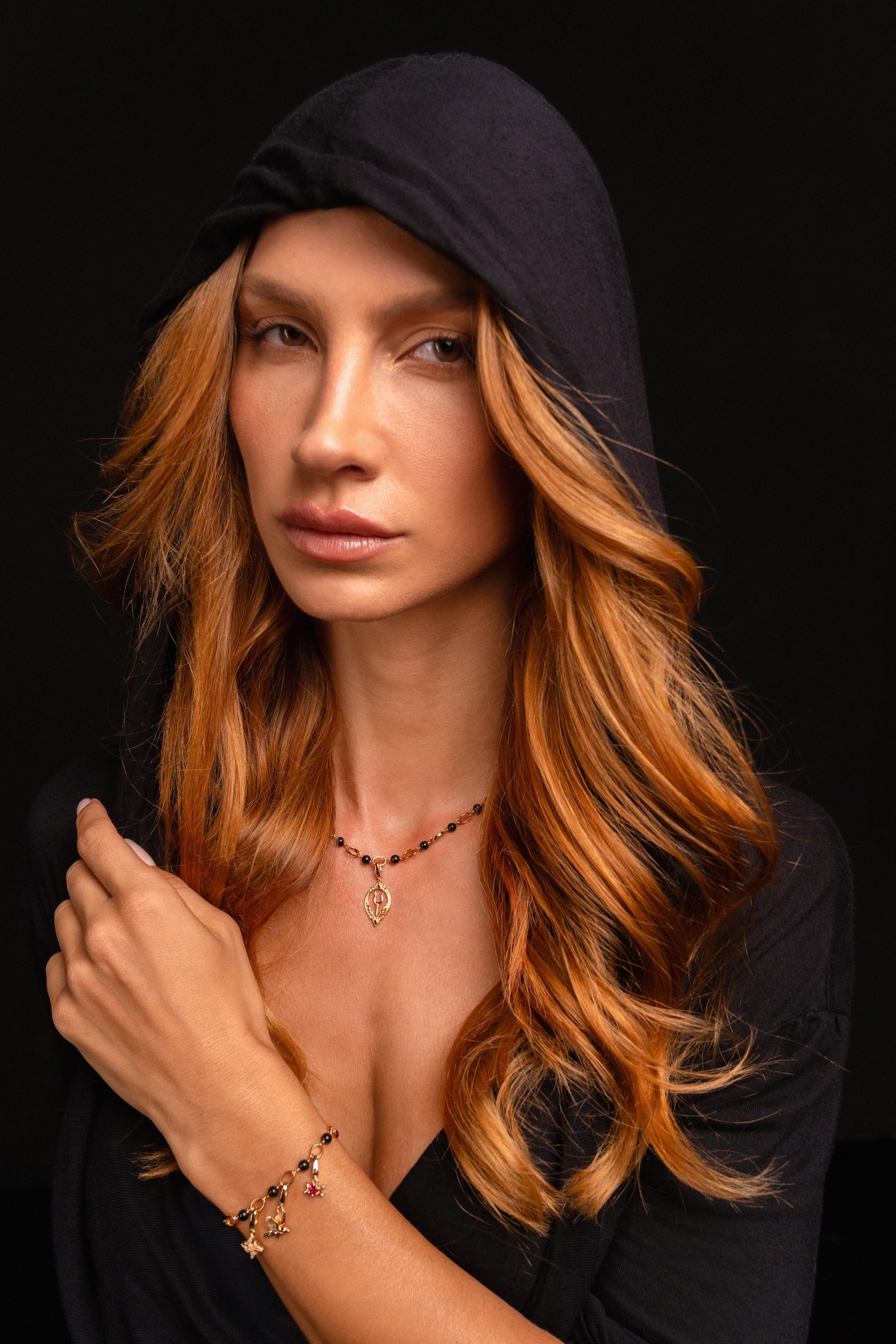 A woman with long, wavy red hair wears a black hooded top and the Preorder: Hades Charm (Ships end October 2026) against a black background, her expression neutral.