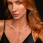 A close-up portrait of a woman with long wavy auburn hair wearing a delicate gold necklace with a small hourglass charm pendant, paired with a black satin camisole against a dark background.