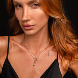 A close-up portrait of a woman with long wavy auburn hair wearing a delicate gold necklace with a small hourglass charm pendant, paired with a black satin camisole against a dark background.
