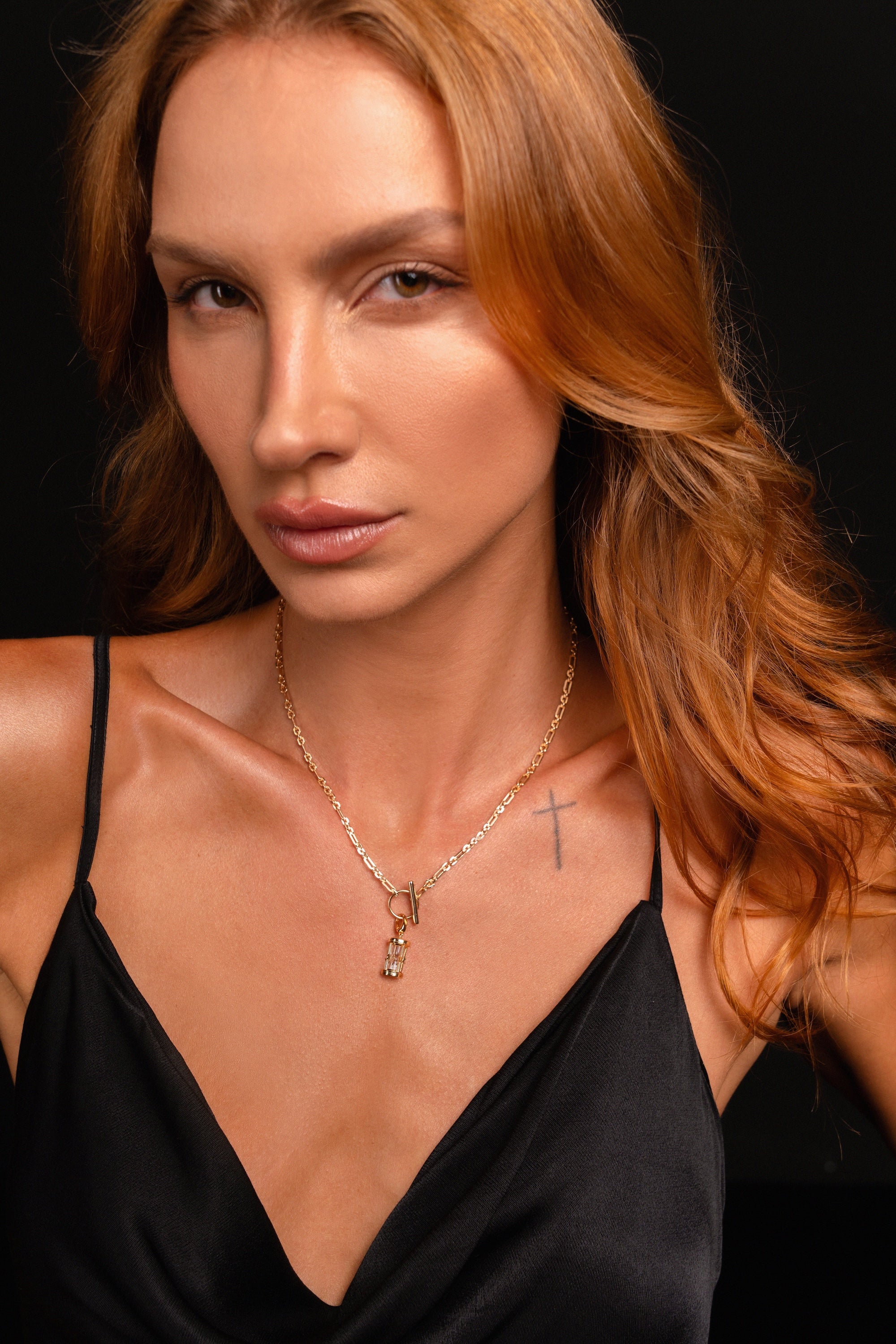 A close-up portrait of a woman with long wavy auburn hair wearing a delicate gold necklace with a small hourglass charm pendant, paired with a black satin camisole against a dark background.