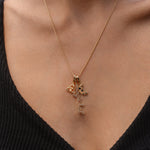 Close-up of a person wearing a gold necklace with three charms: a triquetra symbol, a script ‘magickal’ charm, and a gold crescent moon with black and white stones.