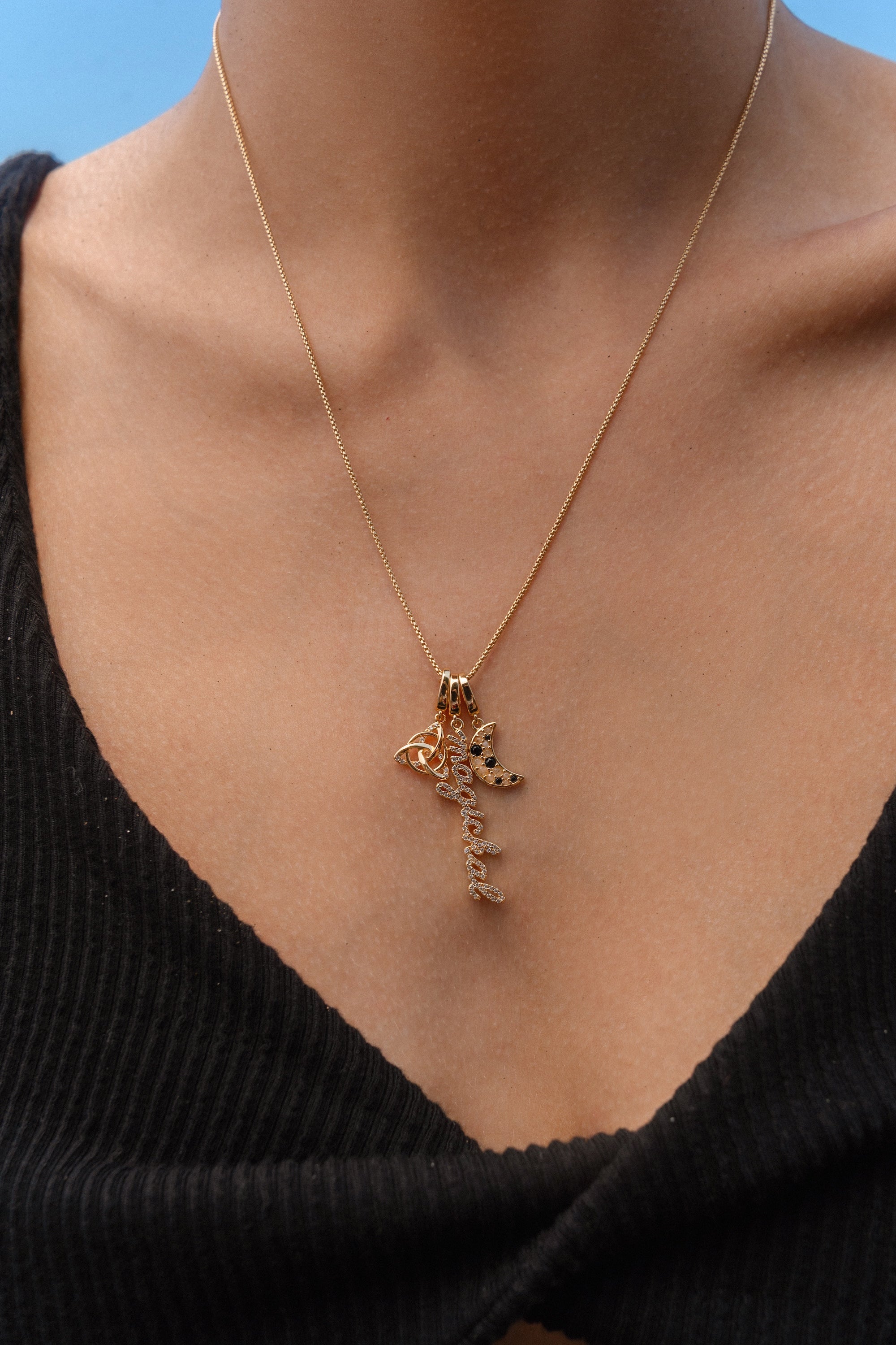 Close-up of a person wearing a gold necklace with three charms: a triquetra symbol, a script ‘magickal’ charm, and a gold crescent moon with black and white stones.