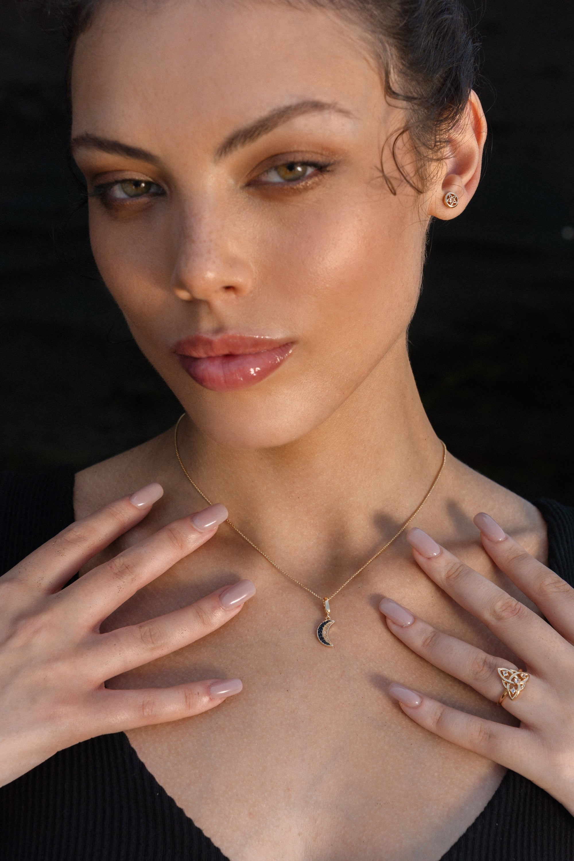 Woman wearing a gold crescent moon necklace, gold pentacle stud earrings, and a gold Triquetra ring with an intricate design.