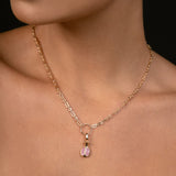 A close-up of a model wearing a delicate rose gold chain necklace with a small pomegranate pendant featuring pink gemstones, set against a dark background.