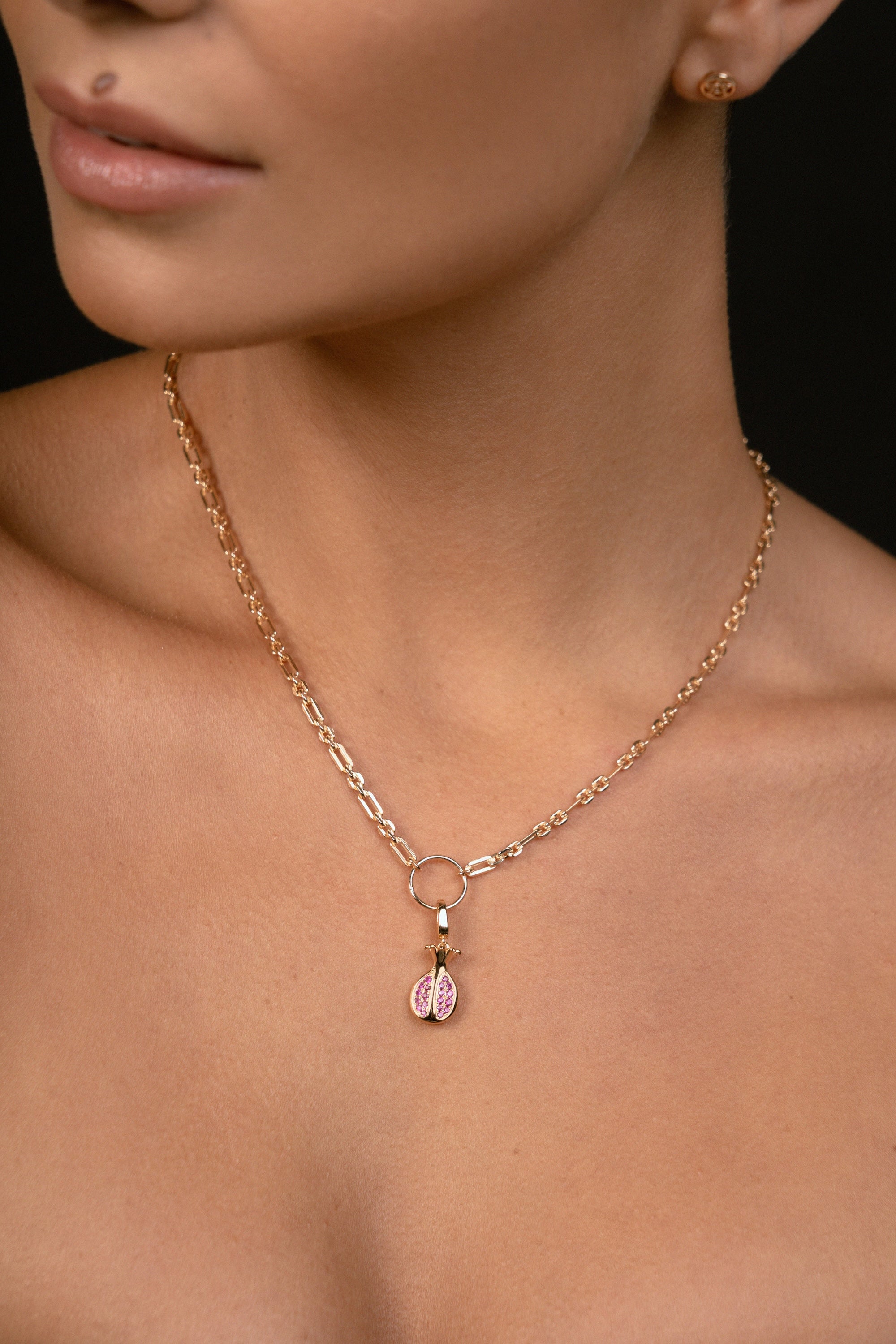 A close-up of a model wearing a delicate rose gold chain necklace with a small pomegranate pendant featuring pink gemstones, set against a dark background.