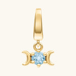 A gold pendant charm featuring a triple moon symbol surrounding a round light blue gemstone, shown on a light cream background.