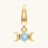 A gold pendant charm featuring a triple moon symbol surrounding a round light blue gemstone, shown on a light cream background.