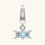 A silver-toned triple moon charm pendant featuring a central light blue round gemstone, set against a solid off-white background.