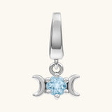 A silver-toned triple moon charm pendant featuring a central light blue round gemstone, set against a solid off-white background.