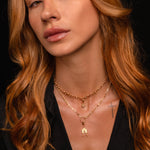 A close-up portrait of a woman with long wavy auburn hair wearing layered gold necklaces, including a Witch's Knot charm pendant and a small house shaped charm, against a dark background.