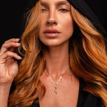 A person with long, wavy reddish hair wears a black hooded garment and the Preorder: Odin Charm (Ships end October 2026) necklace, gazing forward with a neutral expression against a black background.