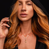 A person with long, wavy reddish hair wears a black hooded garment and the Preorder: Odin Charm (Ships end October 2026) necklace, gazing forward with a neutral expression against a black background.