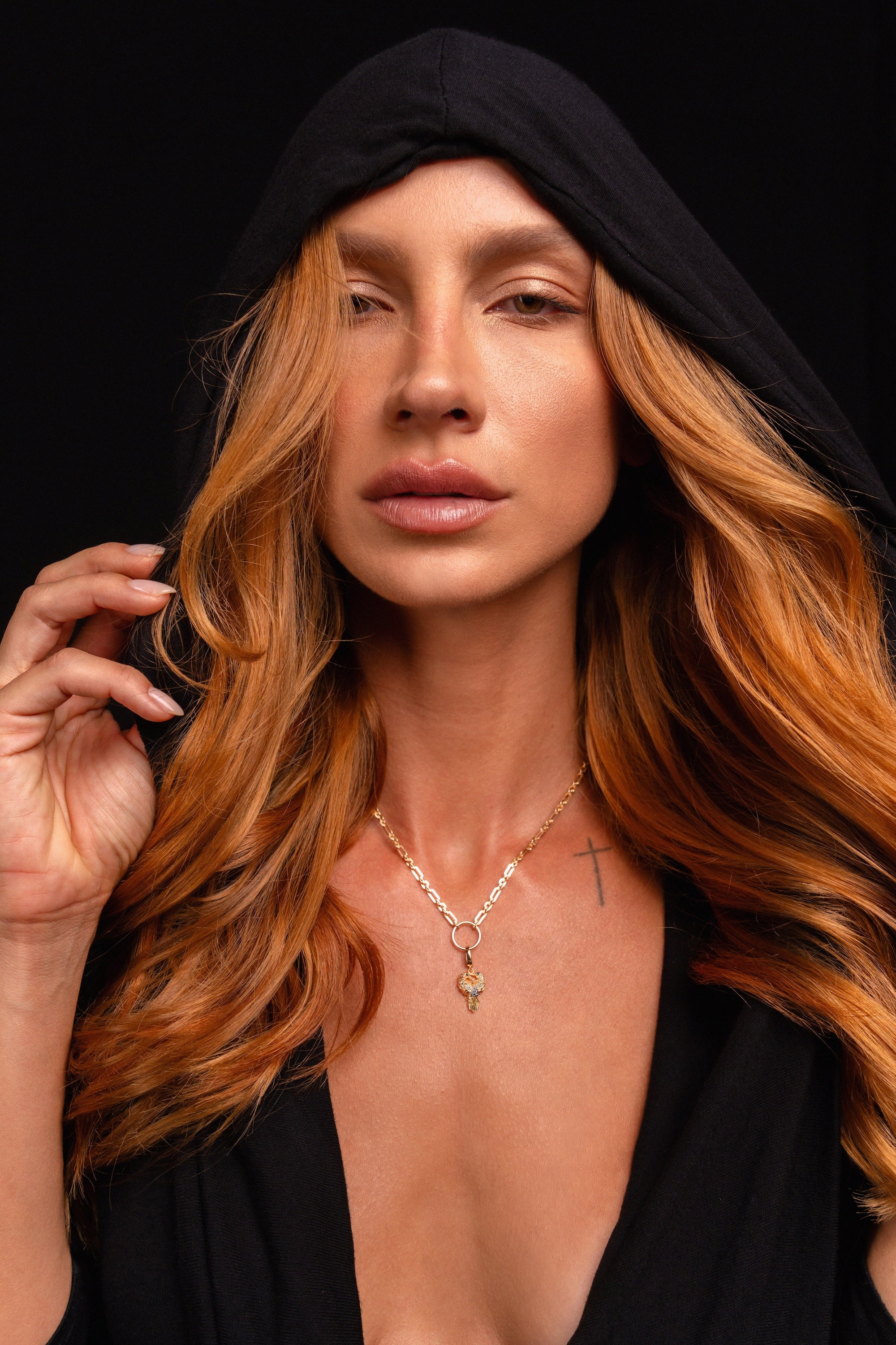A person with long, wavy reddish hair wears a black hooded garment and the Preorder: Odin Charm (Ships end October 2026) necklace, gazing forward with a neutral expression against a black background.