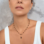 A close-up of a woman wearing a delicate necklace with small black beads and a gold-toned pendant with a dark round stone, paired with a white ribbed tank top against a textured gray background.