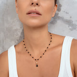 A close-up of a woman wearing a delicate necklace with small black beads and a gold-toned pendant with a dark round stone, paired with a white ribbed tank top against a textured gray background.