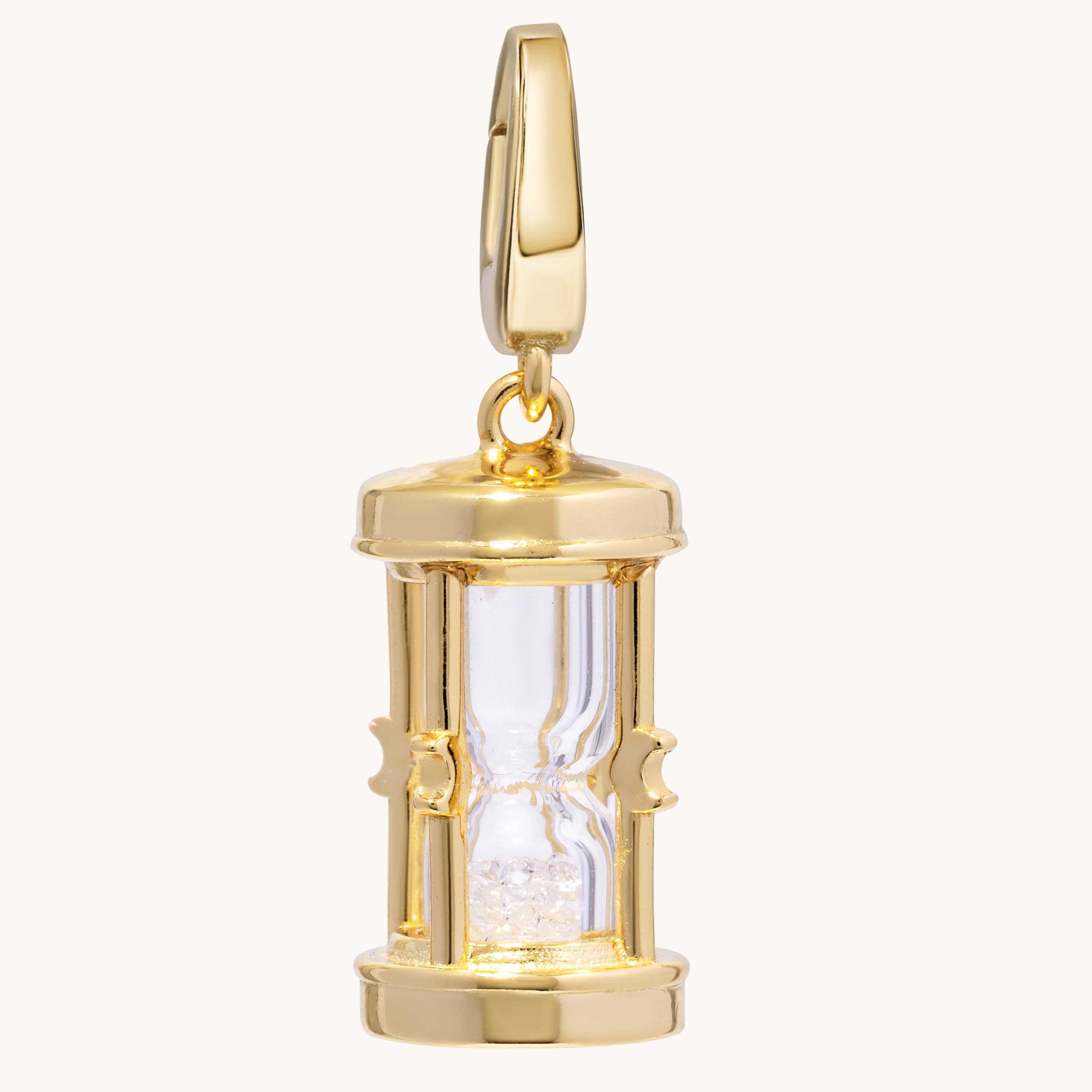 A polished silver hourglass pendant charm with a clear glass center and glittering crystal-toned sand inside, shown on a light cream background.