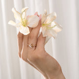 Hand holding two white flowers and wearing a gold moon-shaped ringagainst a light background