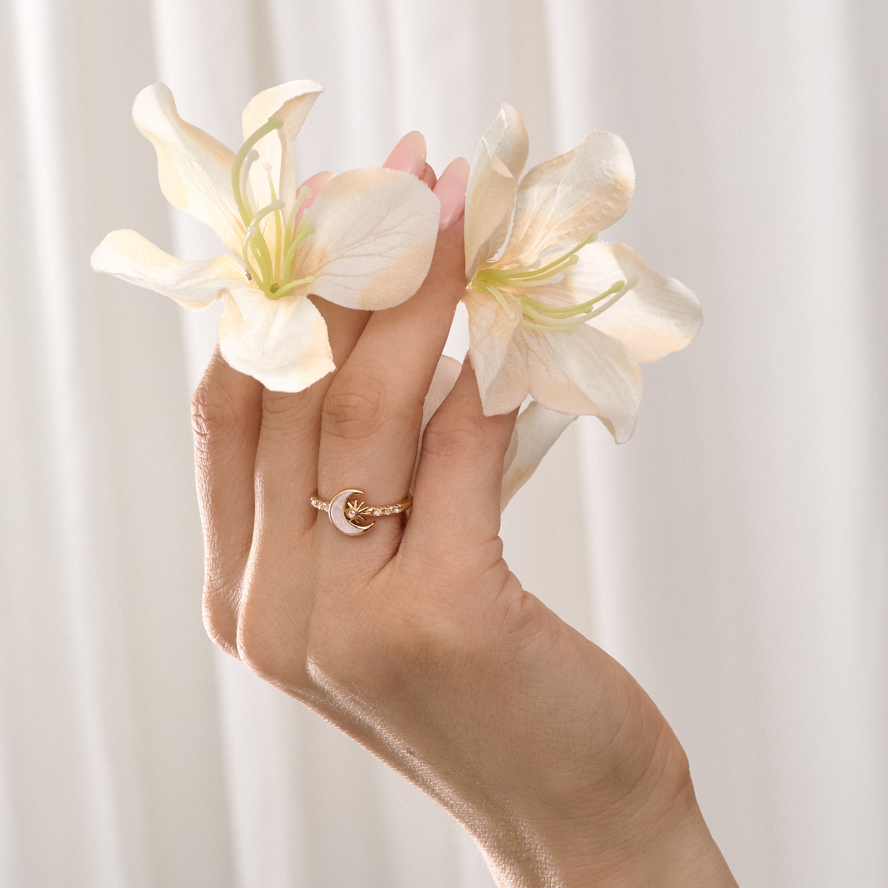 Hand holding two white flowers and wearing a gold moon-shaped ringagainst a light background
