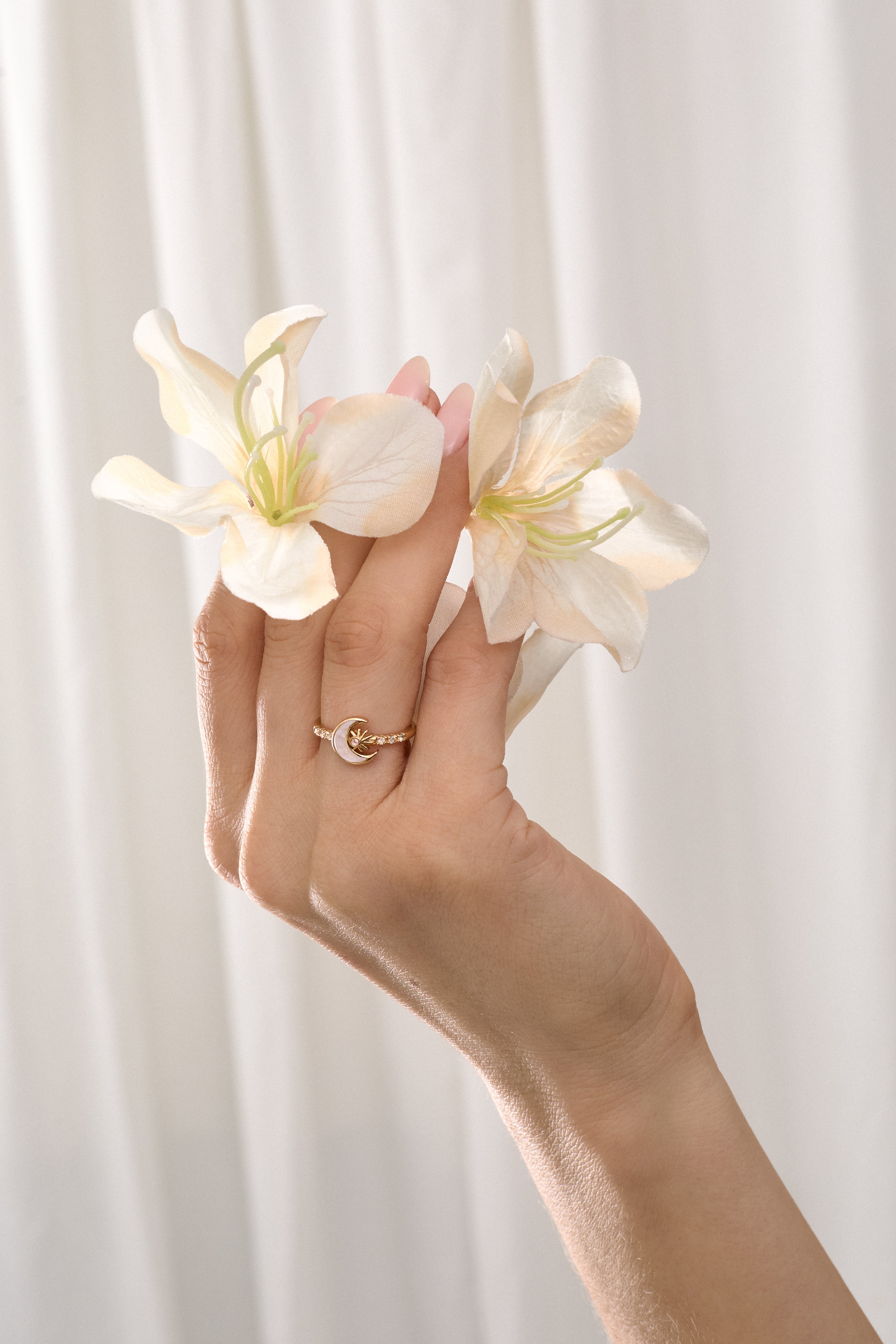 Hand holding two white flowers and wearing a gold moon-shaped ringagainst a light background