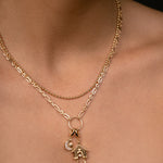 A close-up of a model's neck and chest wearing a delicate gold double-layer necklace. The top layer is a fine chain, and the bottom layer features a textured link chain with a circular ring holding a small house and crescent moon charm.