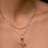 A close-up of a model's neck and chest wearing a delicate gold double-layer necklace. The top layer is a fine chain, and the bottom layer features a textured link chain with a circular ring holding a small house and crescent moon charm.