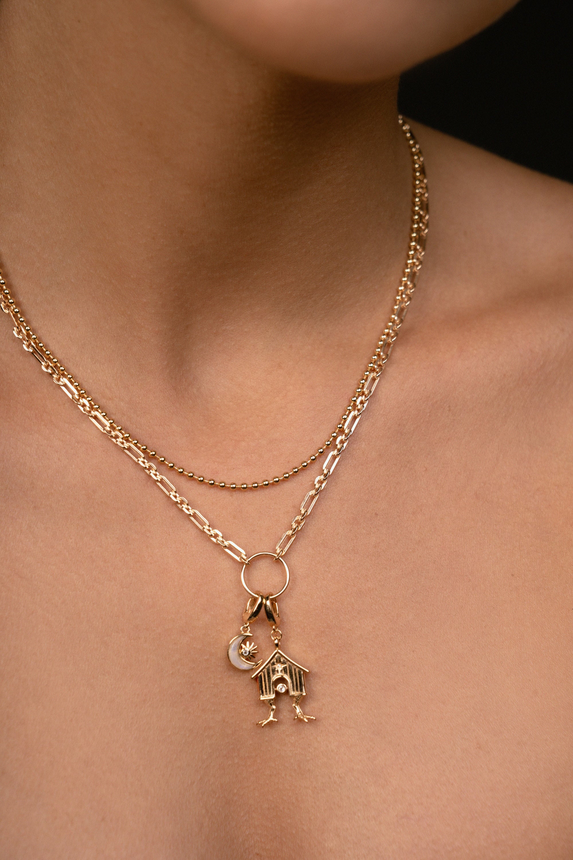 A close-up of a model's neck and chest wearing a delicate gold double-layer necklace. The top layer is a fine chain, and the bottom layer features a textured link chain with a circular ring holding a small house and crescent moon charm.