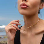 Woman wearing a gold and pearl charm necklace featuring a Hecate’s Wheel charm, a triquetra charm, and a crescent moon charm with black and white stones, photographed by the beach.