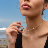 Woman wearing a gold and pearl charm necklace featuring a Hecate’s Wheel charm, a triquetra charm, and a crescent moon charm with black and white stones, photographed by the beach.