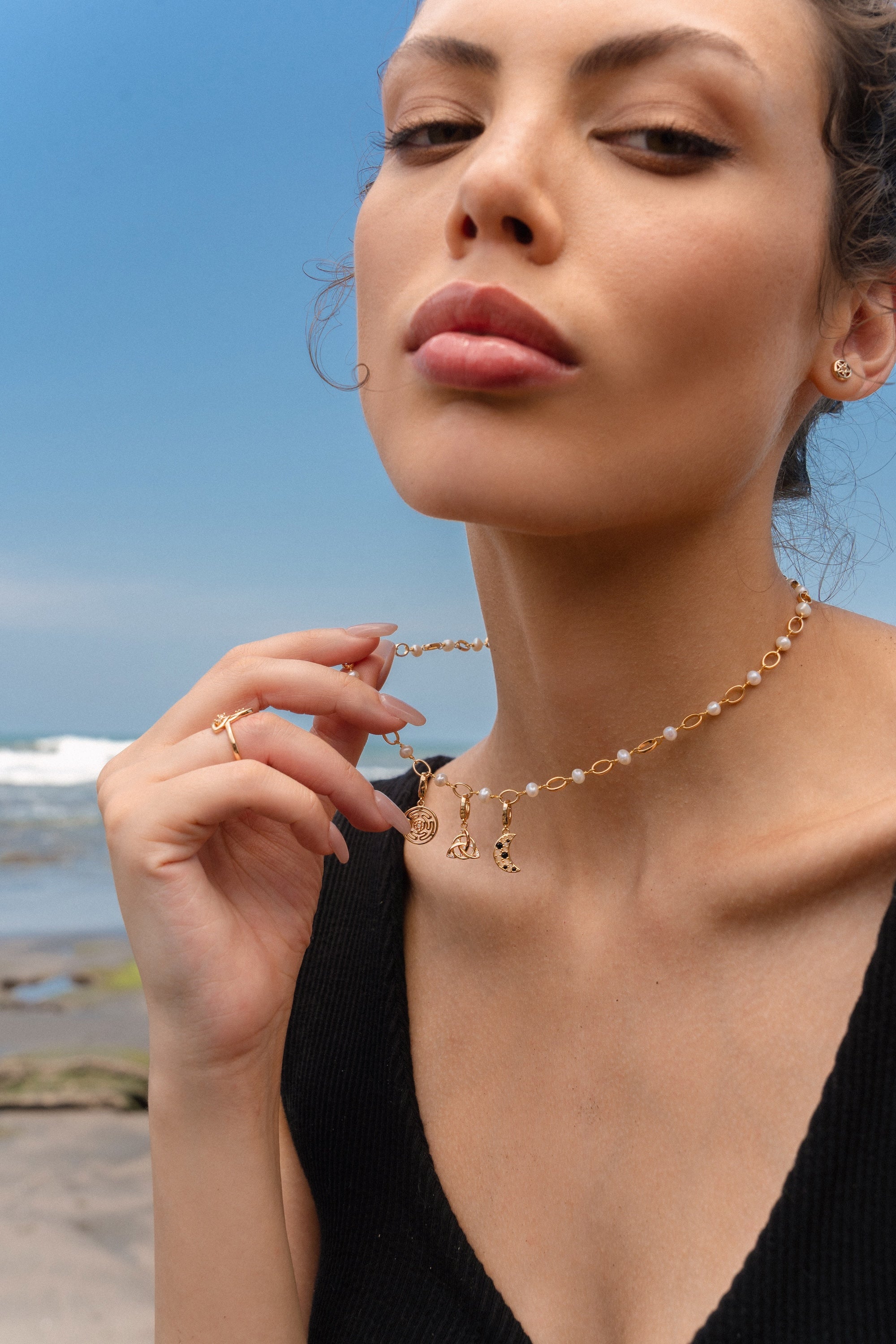 Woman wearing a gold and pearl charm necklace featuring a Hecate’s Wheel charm, a triquetra charm, and a crescent moon charm with black and white stones, photographed by the beach.