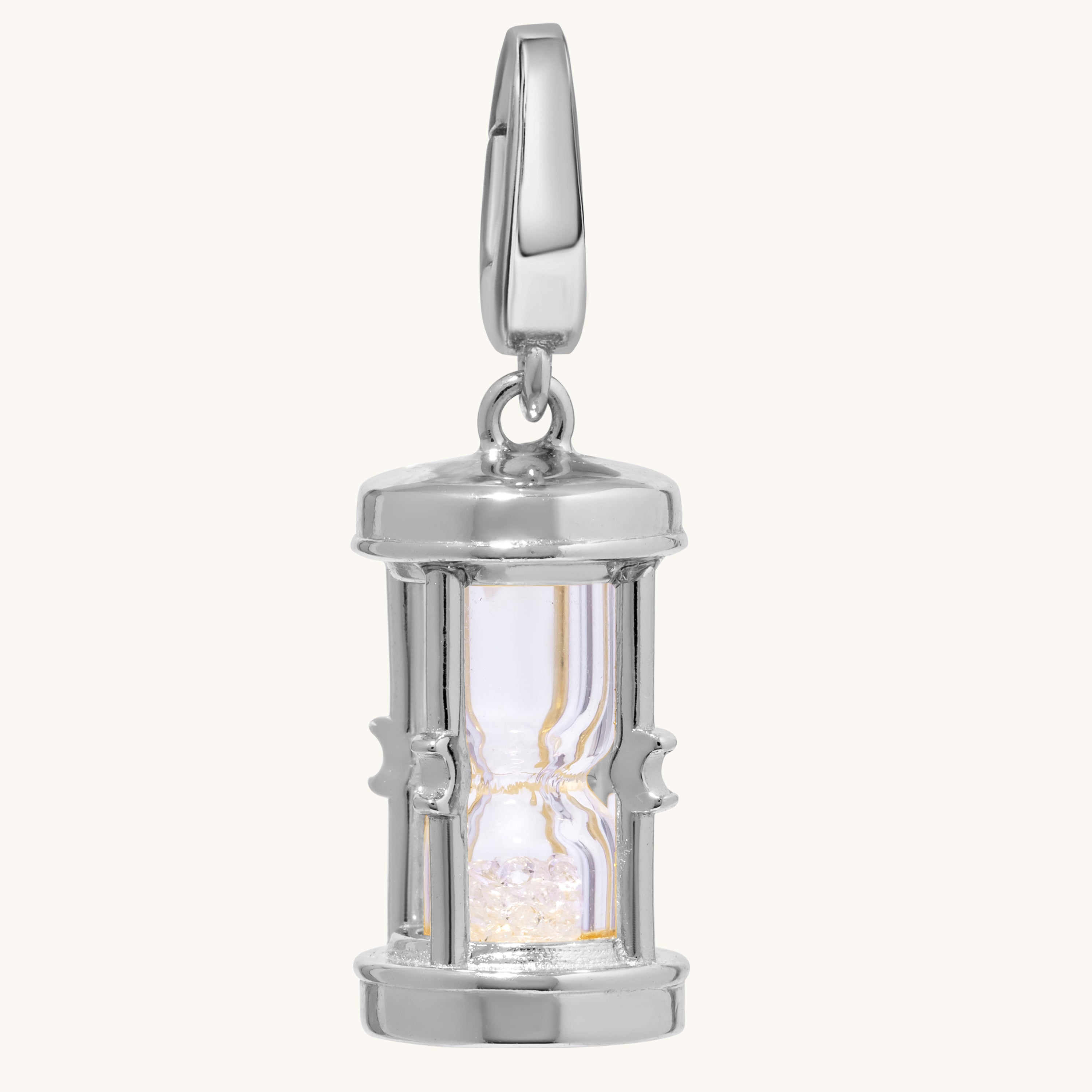 A polished silver hourglass pendant charm with a clear glass center and glittering crystal-toned sand inside, shown on a light cream background.