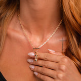 A woman with long, reddish hair wears a gold chain necklace with an hourglass charm pendant and a black top, her manicured hand touching it as a small cross tattoo peeks from her shoulder.