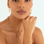 A close-up of a model wearing a gold jewelry set featuring a delicate necklace with small charms, a matching ring, and a bracelet small charms, against a clean white background.
