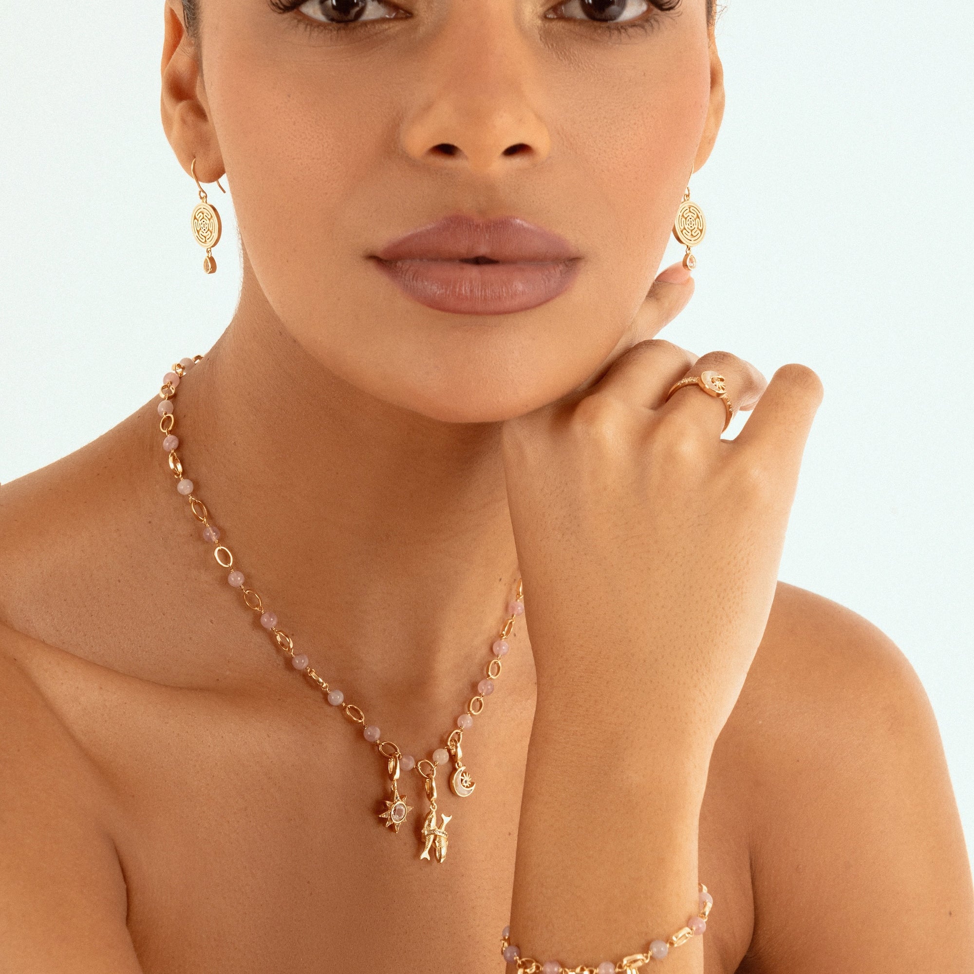 A close-up of a model wearing a gold jewelry set featuring a delicate necklace with small charms, a matching ring, and a bracelet small charms, against a clean white background.