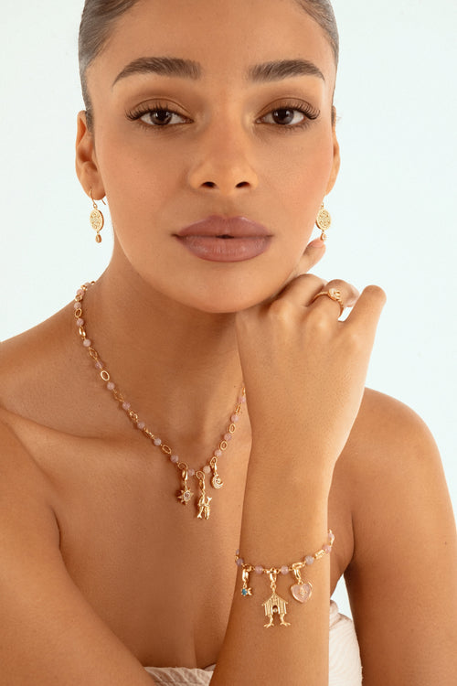 A close-up of a model wearing a gold jewelry set featuring a delicate necklace with small charms, a matching ring, and a bracelet small charms, against a clean white background.