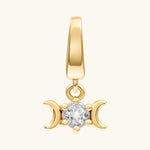 A gold pendant charm featuring a central clear gemstone flanked by two crescent moons, set against a plain off-white background.