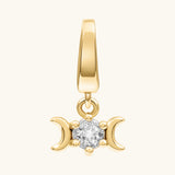 A gold pendant charm featuring a central clear gemstone flanked by two crescent moons, set against a plain off-white background.