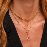 A close-up of a model wearing a layered gold necklace set. The set features a thicker curb chain and a delicate chain with a small house-shaped pendant and a tiny Witch's Knot charm.