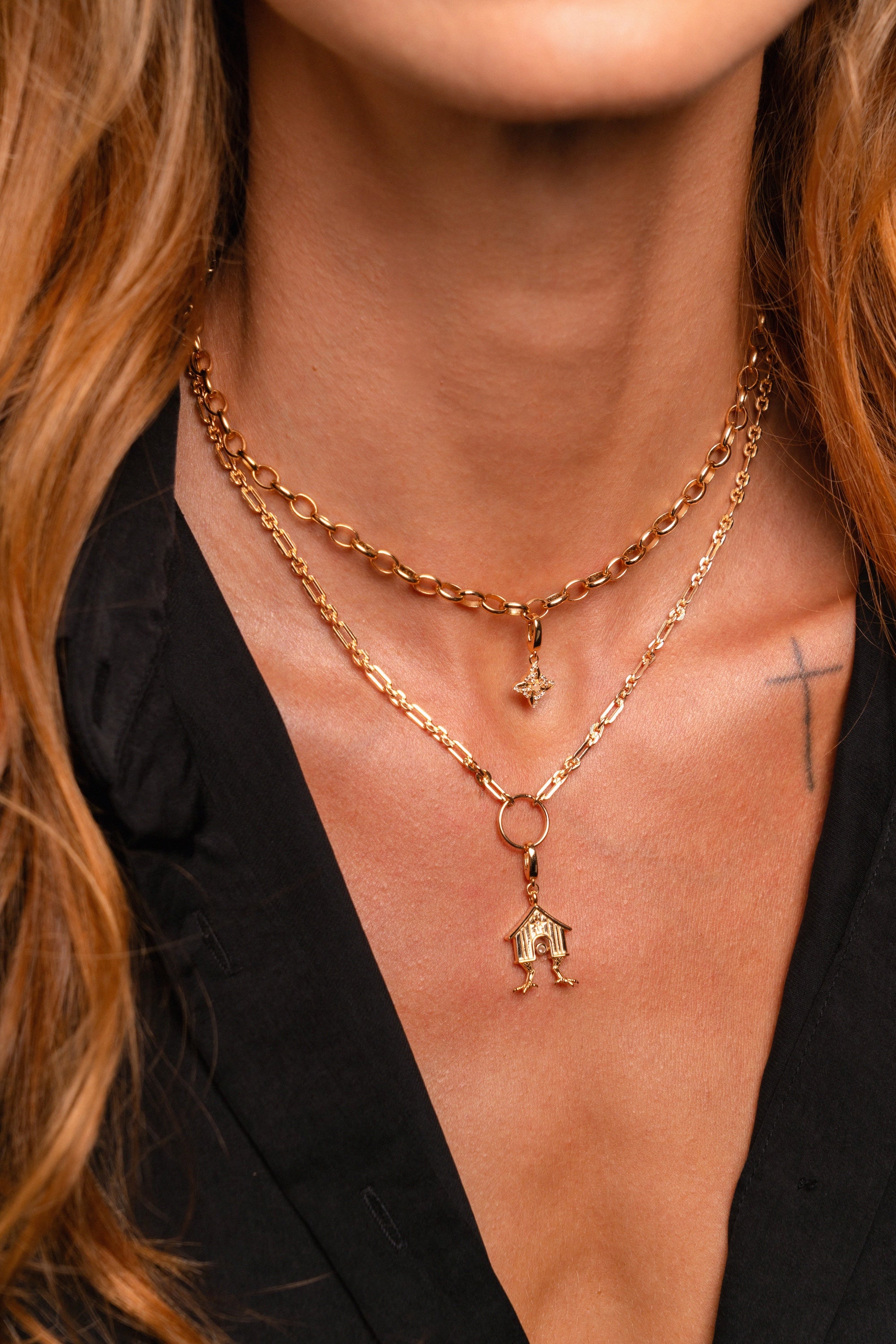 A close-up of a model wearing a layered gold necklace set. The set features a thicker curb chain and a delicate chain with a small house-shaped pendant and a tiny Witch's Knot charm.
