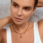 A close-up of a woman with short brown hair wearing a delicate beaded necklace with black and gold beads and a small dark pendant, paired with a white ribbed tank top against a textured gray wall.